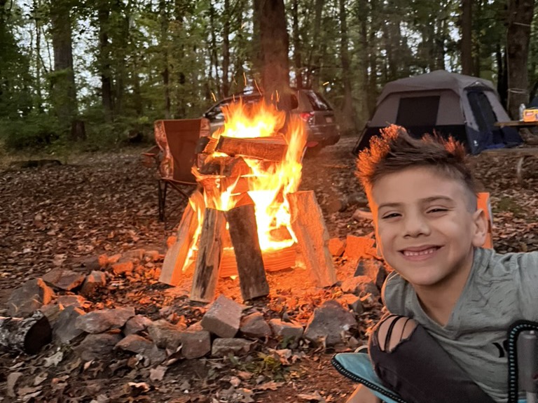 A smiling camper next to a crackling campfire at a wooded fall campsite